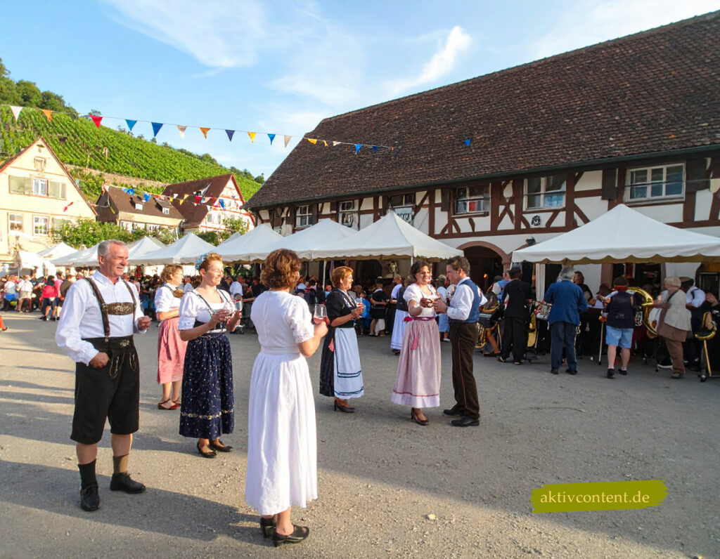 Menschen in regionaler Tracht feiern auf einem Marktplatz in Baden-Württemberg, Deutschland: Fachwerkhäuser, Weinberge, Wimpelketten und Blaskapelle vor Festzelten – stimmungsvolles Fest im Süden der deutschen Bundesländer.
